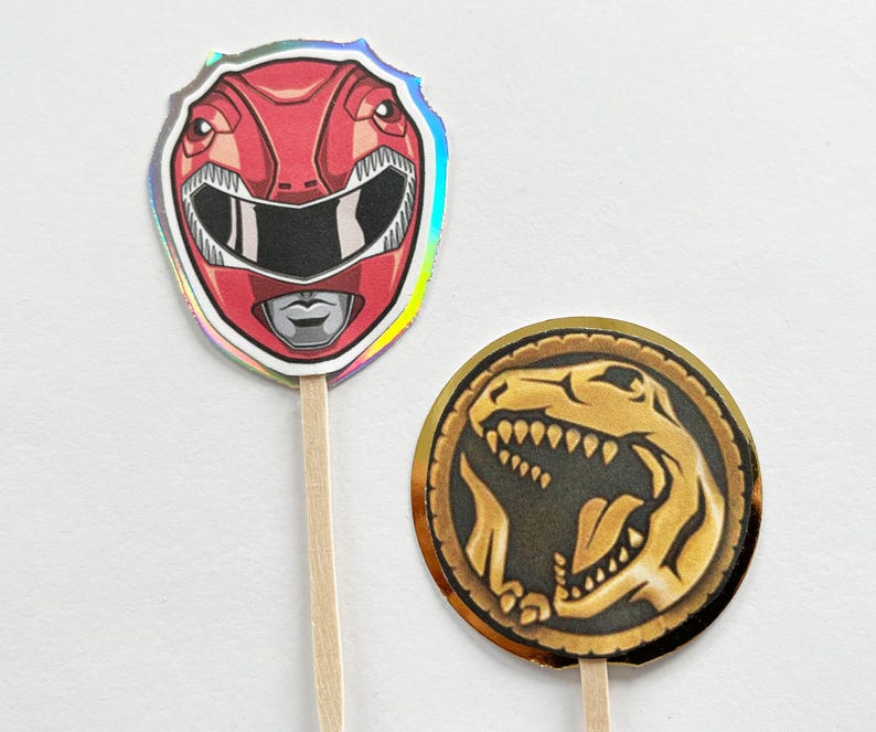 Puede incluir: Dos adornos para cupcakes con tem&aacute;tica de Power Rangers. Uno presenta un casco de Ranger rojo con un contorno plateado. El otro es una moneda de oro con un dise&ntilde;o de dinosaurio negro. Ambos est&aacute;n unidos a palitos de madera.