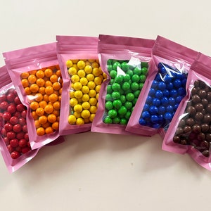 May include: Six small pink resealable bags filled with colorful candy coated chocolate balls. The bags are arranged in a rainbow pattern with red, orange, yellow, green, blue, and brown chocolate balls.