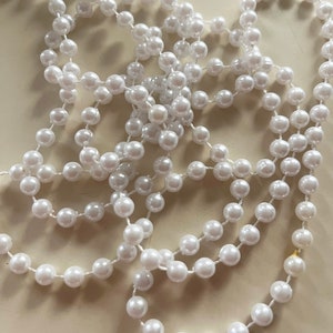 Puede incluir: Una hebra de cuentas de imitación de perlas blancas, enhebradas en un hilo blanco fino. Las cuentas están dispuestas en una pila suelta y enredada.