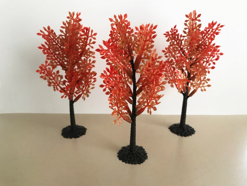 May include: Three small artificial trees with orange leaves and black plastic trunks. The trees are on a light brown surface.