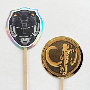 Puede incluir: Dos adornos para cupcakes con tem&aacute;tica de Power Rangers. Uno presenta un casco negro con detalles plateados y un borde hologr&aacute;fico. El otro es una moneda dorada con un dise&ntilde;o de elefante sobre un fondo marr&oacute;n.