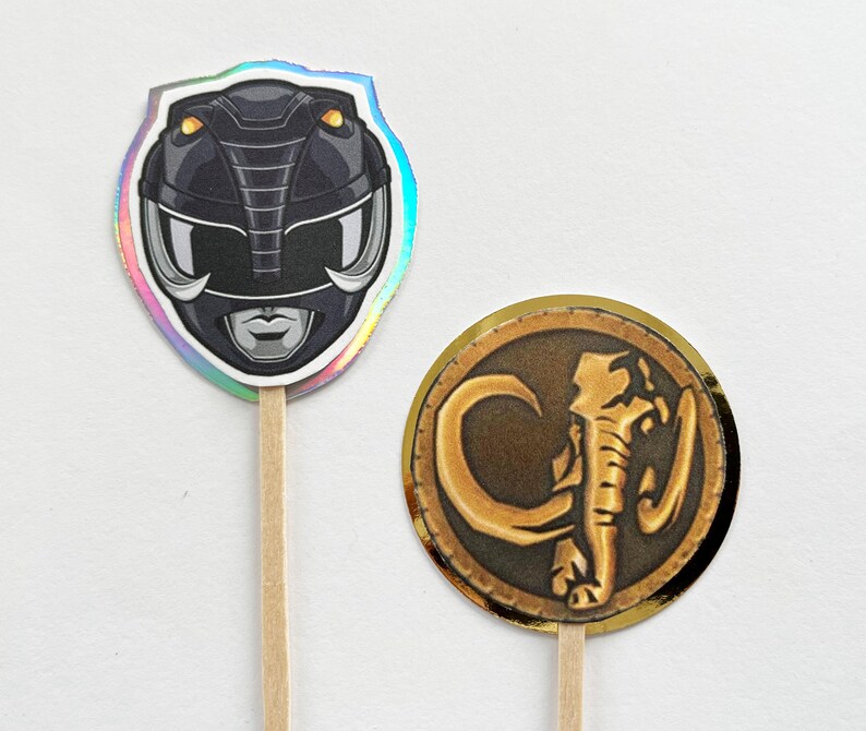 Puede incluir: Dos adornos para cupcakes con tem&aacute;tica de Power Rangers. Uno presenta un casco negro con detalles plateados y un borde hologr&aacute;fico. El otro es una moneda dorada con un dise&ntilde;o de elefante sobre un fondo marr&oacute;n.