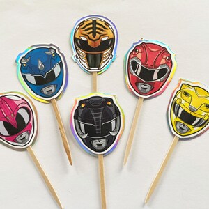 Puede incluir: Una colecci&oacute;n de adornos para cupcakes con tem&aacute;tica de Power Rangers. Cada adorno presenta un dise&ntilde;o de casco de Ranger de diferente color, incluyendo rojo, azul, rosa, amarillo, negro y con rayas de tigre. Los adornos est&aacute;n adheridos a palitos de madera.
