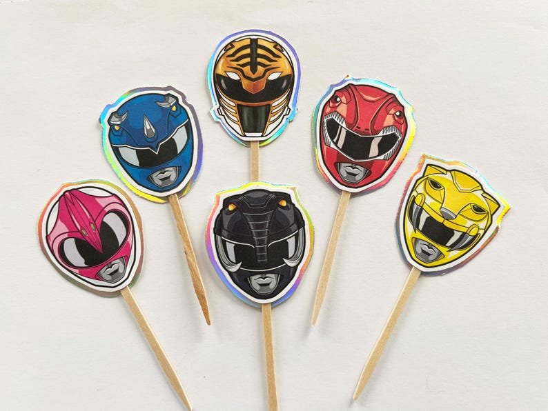 Puede incluir: Una colecci&oacute;n de adornos para cupcakes con tem&aacute;tica de Power Rangers. Cada adorno presenta un dise&ntilde;o de casco de Ranger de diferente color, incluyendo rojo, azul, rosa, amarillo, negro y con rayas de tigre. Los adornos est&aacute;n adheridos a palitos de madera.