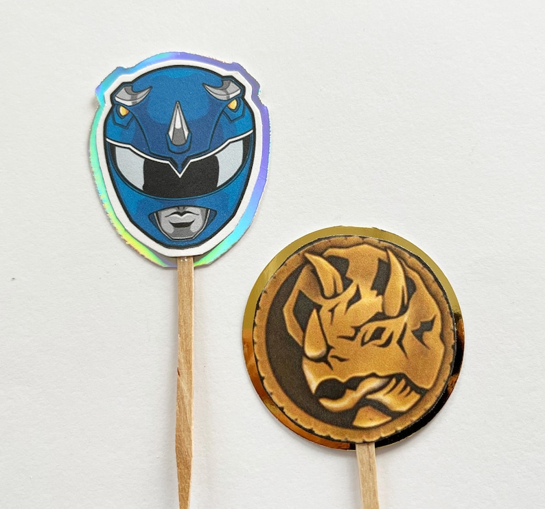 Puede incluir: Dos adornos para cupcakes con tem&aacute;tica de Power Rangers. Uno es un casco de Ranger azul con un contorno plateado y un borde hologr&aacute;fico. El otro es una moneda de oro con un dise&ntilde;o de rinoceronte. Ambos est&aacute;n unidos a palitos de madera.