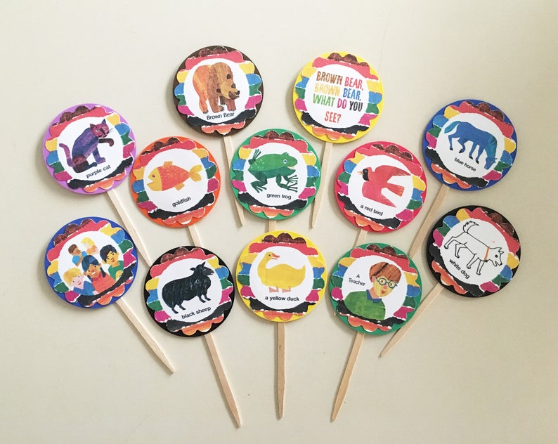 May include: Brown Bear, Brown Bear, What Do You See? themed cupcake toppers. Each topper features a colorful circle with an illustration and text from the book. The toppers include a purple cat, a goldfish, a green frog, a red bird, a blue horse, a white dog, a group of children, a black sheep, a yellow duck, and a teacher.