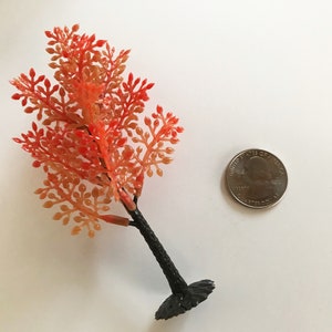 May include: A small, artificial autumn tree with orange and red leaves. The tree is about the size of a 25 pence coin.