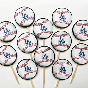May include: Baseball-themed cupcake toppers featuring a baseball design with red stitching and the letters "LA" in blue. Each topper is attached to a wooden stick. Perfect for a baseball-themed party or event.