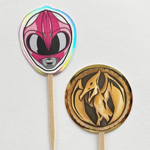 Puede incluir: Dos adornos para cupcakes con tem&aacute;tica de Power Rangers. Uno presenta un casco rosa con un borde hologr&aacute;fico, el otro una moneda de oro con un emblema de dinosaurio. Ambos est&aacute;n unidos a palitos de madera.