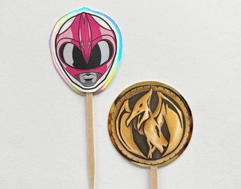 Puede incluir: Dos adornos para cupcakes con tem&aacute;tica de Power Rangers. Uno presenta un casco rosa con un borde hologr&aacute;fico, el otro una moneda de oro con un emblema de dinosaurio. Ambos est&aacute;n unidos a palitos de madera.