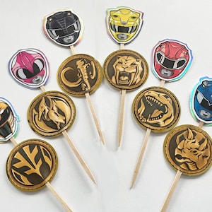 Puede incluir: Adornos para cupcakes con tem&aacute;tica de Power Rangers. Los adornos presentan varios cascos de Power Rangers en colores como rosa, amarillo, rojo y azul, junto con medallones dorados con dise&ntilde;os de animales. Cada adorno est&aacute; unido a un palillo de madera.