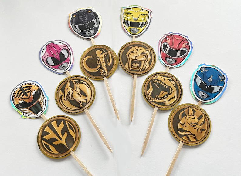Puede incluir: Adornos para cupcakes con tem&aacute;tica de Power Rangers. Los adornos presentan varios cascos de Power Rangers en colores como rosa, amarillo, rojo y azul, junto con medallones dorados con dise&ntilde;os de animales. Cada adorno est&aacute; unido a un palillo de madera.