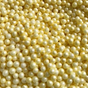 May include: A close-up image of a large quantity of small, round, pearl-like sprinkles. The sprinkles are a pale yellow color.