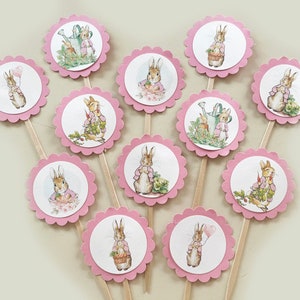 May include: Twelve pink cupcake toppers with scalloped edges feature illustrations of Peter Rabbit in various poses. Each topper has a wooden stick for inserting into cupcakes.