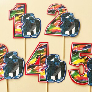 May include: Five colorful cake toppers with a gorilla and a number on each. The numbers are 1, 2, 3, 4, and 5. The gorillas are blue and gray and have a white background. The numbers are on a white background with colorful brushstrokes.