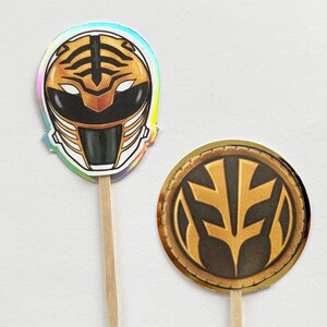 Puede incluir: Dos adornos para cupcakes con tem&aacute;tica de Power Rangers. Uno presenta un casco blanco y dorado con detalles negros y un dise&ntilde;o de rayas de tigre. El otro es un emblema circular dorado y negro. Ambos est&aacute;n unidos a palitos de madera.