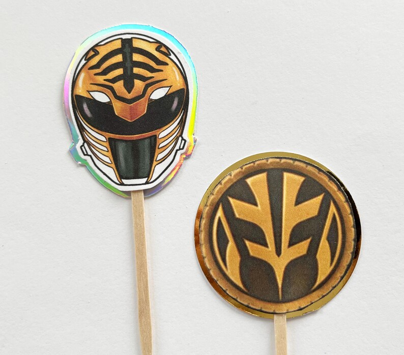 Puede incluir: Dos adornos para cupcakes con tem&aacute;tica de Power Rangers. Uno presenta un casco blanco y dorado con detalles negros y un dise&ntilde;o de rayas de tigre. El otro es un emblema circular dorado y negro. Ambos est&aacute;n unidos a palitos de madera.