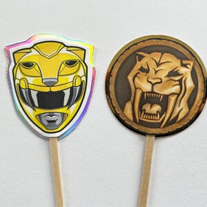 Puede incluir: Dos adornos para cupcakes con tem&aacute;tica de Power Rangers. Uno es un casco de Ranger amarillo con un borde hologr&aacute;fico. El otro es un emblema circular con un dise&ntilde;o de tigre dientes de sable en tonos marrones y dorados.