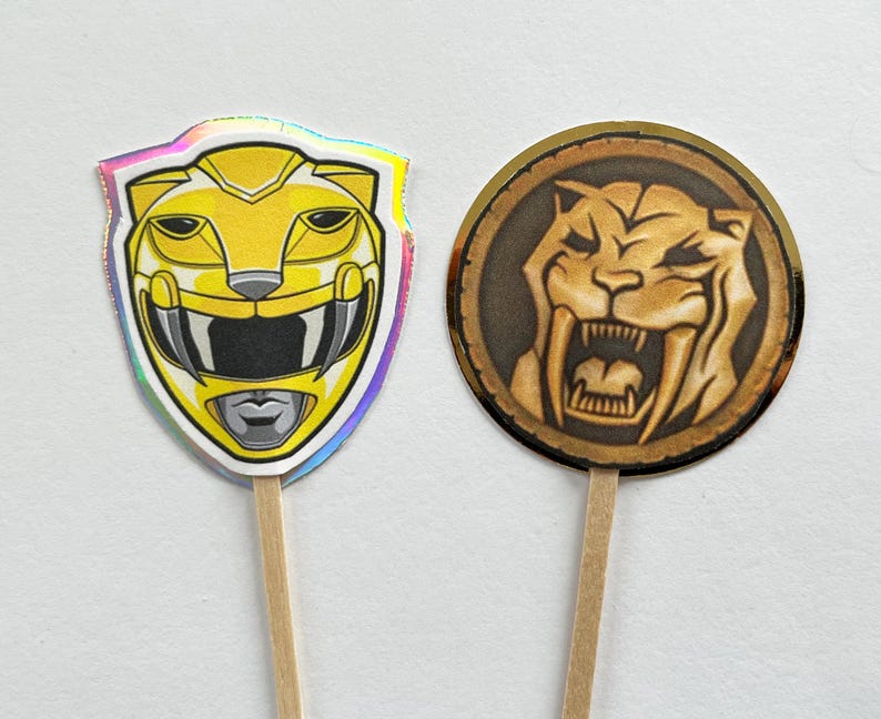 Puede incluir: Dos adornos para cupcakes con tem&aacute;tica de Power Rangers. Uno es un casco de Ranger amarillo con un borde hologr&aacute;fico. El otro es un emblema circular con un dise&ntilde;o de tigre dientes de sable en tonos marrones y dorados.