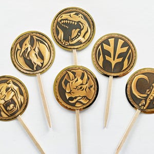 Puede incluir: Conjunto de siete adornos para cupcakes de Power Rangers dorados y bronce. Cada adorno circular muestra un animal o s&iacute;mbolo diferente, como un T-Rex o un elefante, fijado a un palillo de madera.