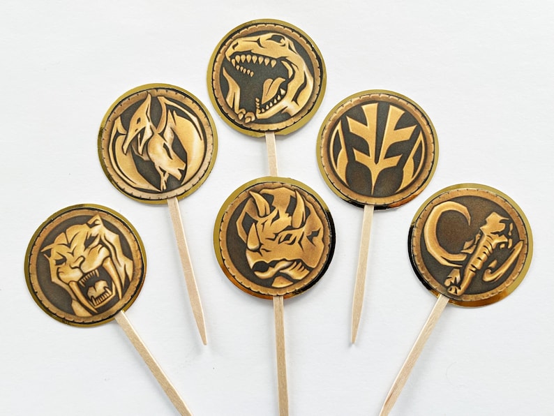 Puede incluir: Conjunto de siete adornos para cupcakes de Power Rangers dorados y bronce. Cada adorno circular muestra un animal o s&iacute;mbolo diferente, como un T-Rex o un elefante, fijado a un palillo de madera.
