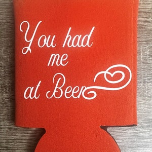 Pode incluir: Resfriador de latas de espuma vermelha com o texto "You had me at Beer" em fonte cursiva branca.