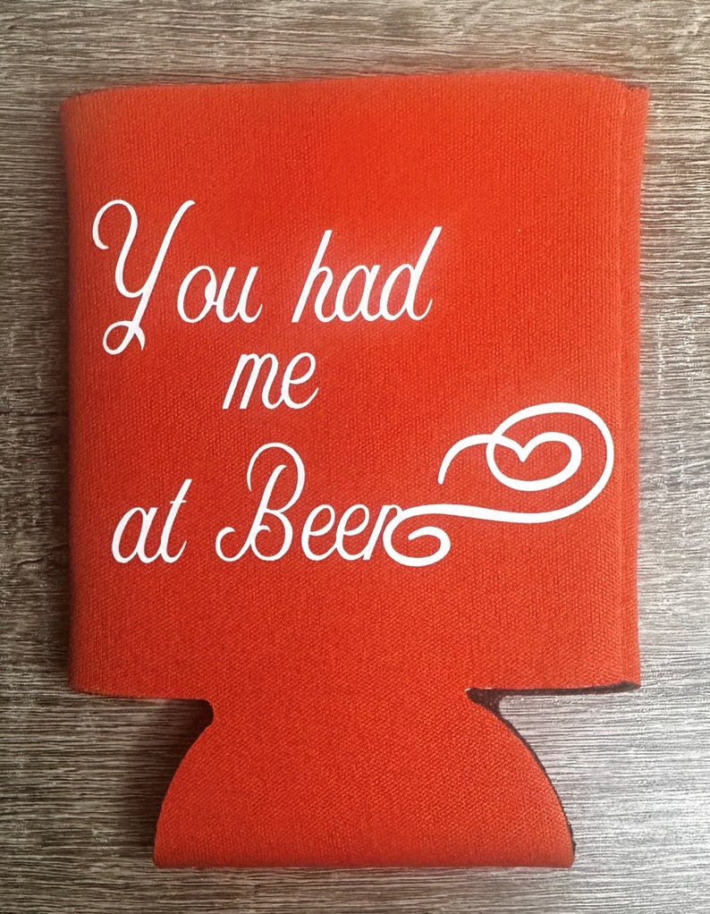 Pode incluir: Resfriador de latas de espuma vermelha com o texto "You had me at Beer" em fonte cursiva branca.