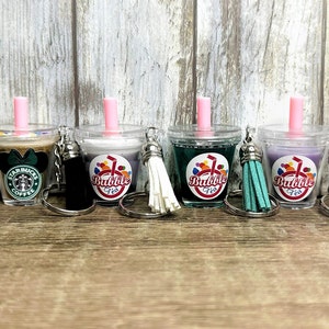 May include: Five clear plastic keychain charms shaped like bubble tea cups with pink straws. The charms have different colored liquids and logos. One charm has a Starbucks logo and a Mickey Mouse bow. The other four charms have a logo that says "Bubble Tea".