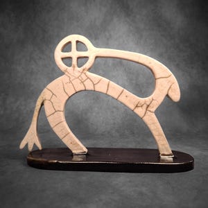 Könnte beinhalten: Eine weiße Keramikskulptur eines stilisierten Tieres mit einem kreisförmigen Design auf dem Kopf. Die Skulptur steht auf einem schwarzen Sockel.