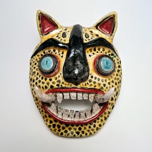 Könnte beinhalten: Eine Keramikmaske mit einem Jaguar-Design. Die Maske hat eine gelbe Basis mit schwarzen Flecken, schwarzen Augenbrauen, einer schwarzen Nase und blauen Augen. Die Ohren sind rot und der Mund hat weiße Zähne und rotes Zahnfleisch.