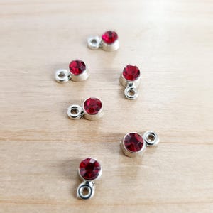 Könnte beinhalten: Sechs rote Strass-Anhänger mit silbernen Ösen. Die Strasssteine sind rund und haben ein facettierten Design.