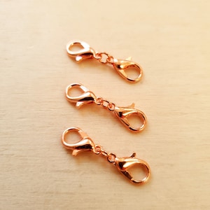 May include: Four gold-toned metal lobster clasps, also known as trigger clasps, are shown on a light brown wooden surface. These clasps are commonly used for jewelry making and crafting.