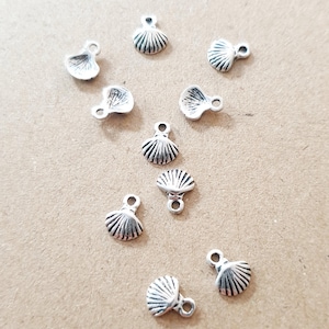 Könnte beinhalten: Eine Sammlung kleiner, silberfarbener Muschelanhänger. Jeder Anhänger hat ein detailliertes, geriffeltes Design und eine kleine Schlaufe zum Befestigen an Schmuck oder anderen Bastelarbeiten. Die Anhänger sind auf einer neutral getönten Oberfläche verstreut.