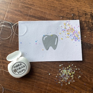 May include: A white container of dental floss labeled "Magic Fairy Rope" next to a white card with a gray tooth graphic and scattered glitter. The floss extends from the container to the card.