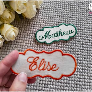 Peut inclure: Deux patchs brodés avec des noms, l'un avec le nom "Matthew" en fil vert sur fond blanc et l'autre avec le nom "Elise" en fil orange sur fond blanc.