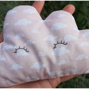 Può includere: Un cuscino a forma di nuvola rosa con nuvole bianche e ciglia ricamate. Il cuscino è fatto a mano e ha il testo "Gostli handmade with love" sul lato destro.
