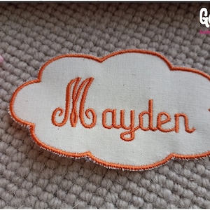 Puede incluir: Un parche de tela blanco en forma de nube con un nombre bordado en naranja "Mayden" en él.
