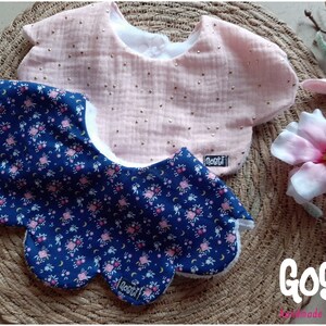 Puede incluir: Dos baberos para bebé. Uno es rosa claro con puntos dorados, el otro es azul marino con un estampado floral. Ambos tienen un reverso blanco y están sobre una esterilla tejida. Los baberos tienen la marca "Gosti".