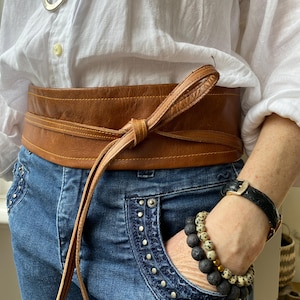 May include: A wide, tan leather belt tied at the front, worn over a white shirt and blue jeans. The belt has a tie closure. The jeans have decorative studs around the pockets. A black watch and beaded bracelets are visible.
