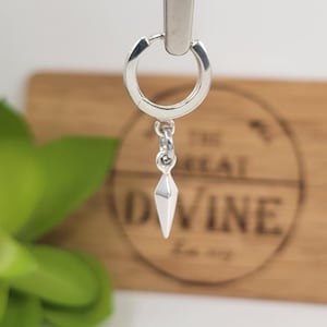 May include: A silver hoop earring with a geometric, diamond-shaped charm. The earring is suspended from a hinged hoop, showcasing a polished, reflective surface. The charm is a faceted, pointed design, adding a modern touch to the jewelry.
