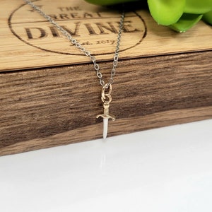 May include: A silver chain necklace with a gold dagger pendant. The pendant is a simple design with a pointed tip and a rounded handle.