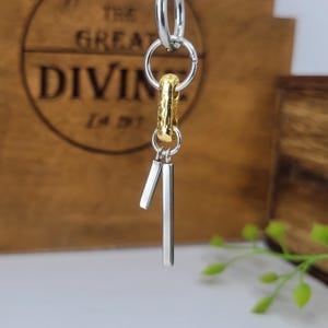 May include: A silver-toned metal pendant featuring a series of interconnected circular rings, a single textured gold-toned oval link, and two smooth silver-toned vertical bar charms of different lengths hanging from the bottom of the structure.