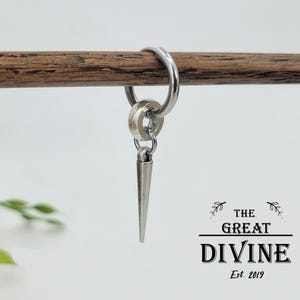 May include: A silver hoop earring with a small, cone-shaped charm. The earring is suspended from a wooden dowel. The Great Divine Est. 2019 logo is in the lower right corner.