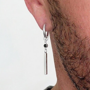 May include: A silver hoop earring with a dangling rectangular bar and a small black gem. The earring is worn in the ear and hangs down. The background is a plain white.
