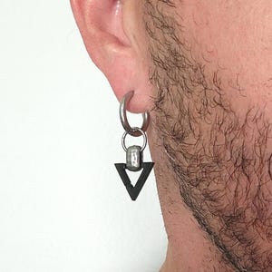 Op de afbeelding: Een zilveren oorring met een zwarte driehoekhanger. De oorring is bevestigd aan een kleine zilveren ring en een rechthoekig zilveren stuk. De driehoek is effen zwart.