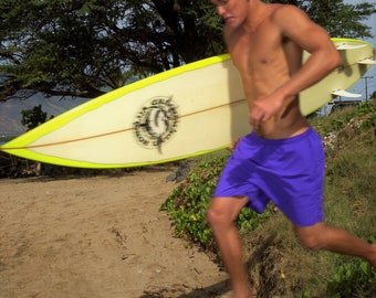 May include: A young man in purple swim trunks holds a yellow surfboard with a black logo. He is standing on a sandy beach with green foliage in the background.