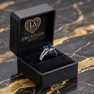 以下が含まれることがあります： 金色の「IFRA ATELIER JEWELRY」の文字とハート型のロゴが入った黒い革製のジュエリーボックス。内部には、大きな正方形の青い宝石が付いたシルバーリングが展示されています。リングは、小さな透明な石が付いたダブルバンドです。