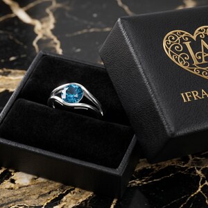 May include: A silver ring with a large, round, blue gemstone sits in an open black jewelry box. The box is next to a closed black box with gold lettering and a heart-shaped logo that reads "IFRA ATELIER JEWELRY". The background is a black and gold marble surface.