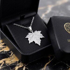 May include: A silver maple leaf pendant necklace displayed in a black jewelry box. The box is open, revealing the necklace with a detailed leaf pendant. A second box with a gold heart logo and the text "IFRA ATELIER JEWELRY" is next to it.