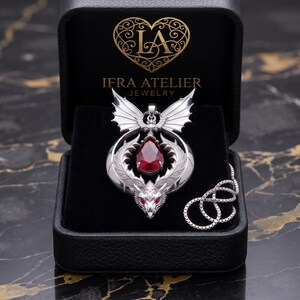 Könnte beinhalten: Ein silberfarbener Drachenanhänger mit einem birnenförmigen roten Edelstein, präsentiert in einer schwarzen Schmuckschatulle mit dem Text IFRA ATELIER JEWELRY und einem Herzlogo. Der Anhänger enthält eine passende silberne Kette.